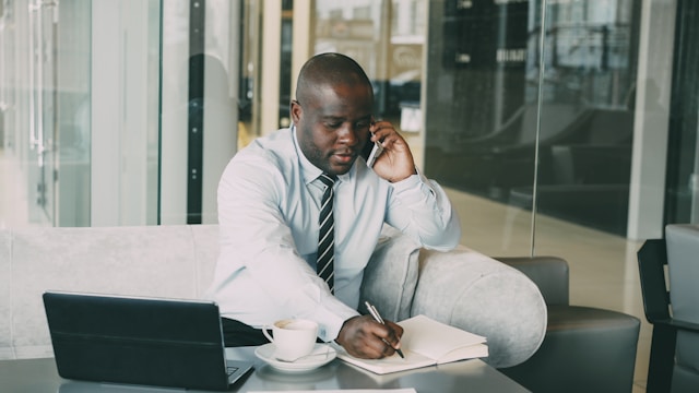 Man sitting in a café talking on the phone while writing in a notebook and working on a laptop.